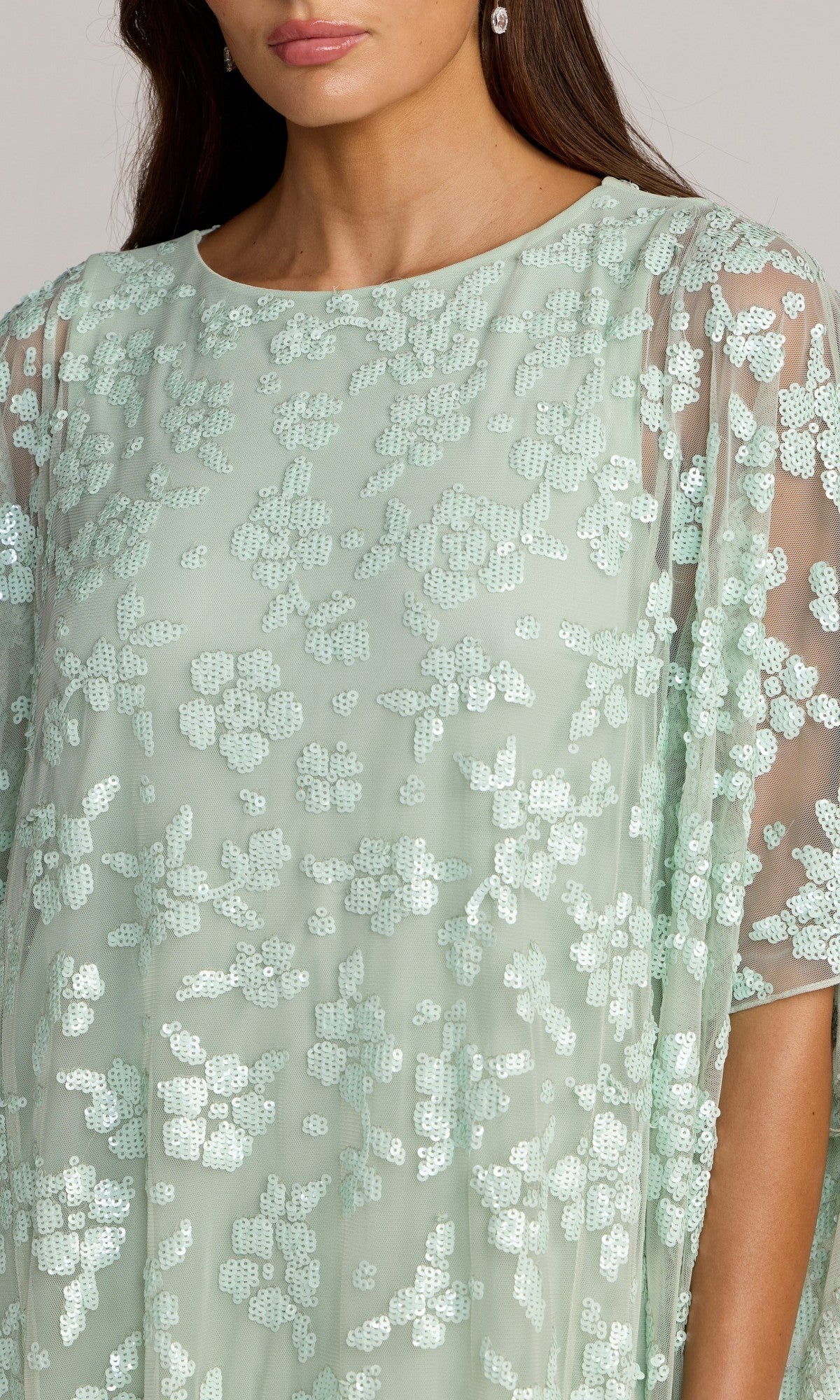 A woman wearing the Mac Duggal 9441 long formal dress in mint green floral sequins, featuring sheer cape sleeves; only her lower face and upper body are visible.